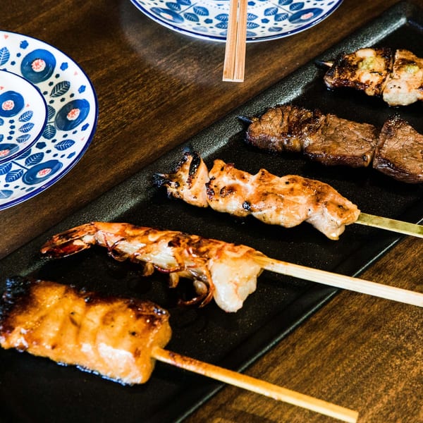 Grilled skewers of shrimp, chicken, and beef yakitori on a black plate with patterned dishes.