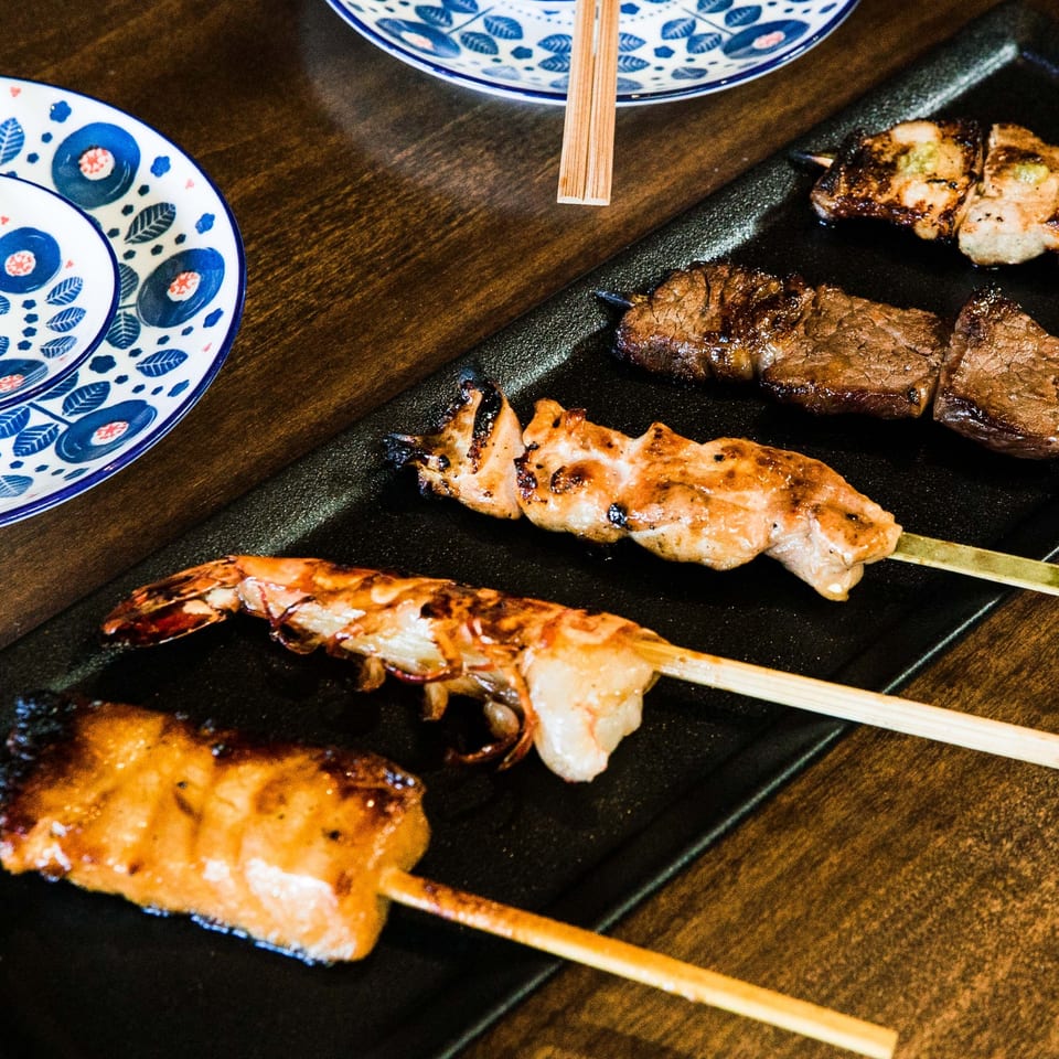 Grilled skewers of shrimp, chicken, and beef yakitori on a black plate with patterned dishes.