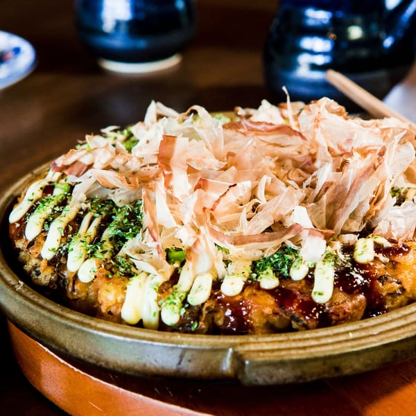 Okonomiyaki topped with bonito flakes, mayonnaise, and green herbs on a brown plate.