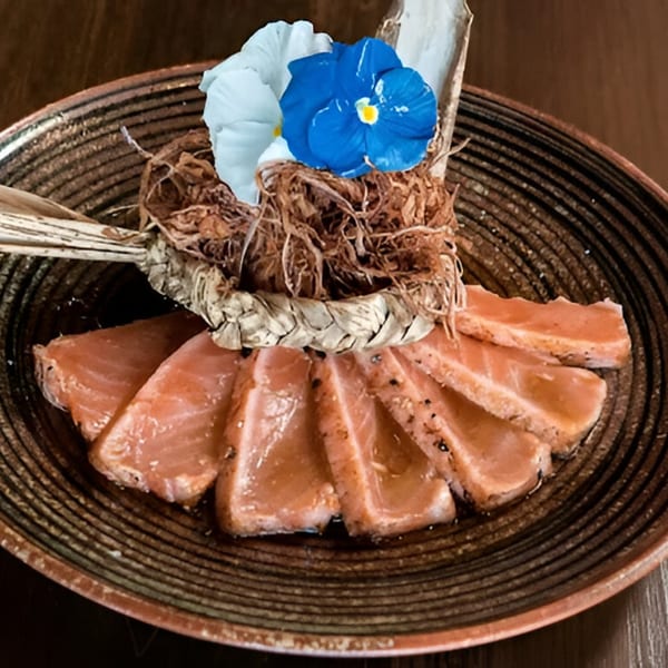 Seared salmon sashimi slices on a dark plate, garnished with blue and white flowers.