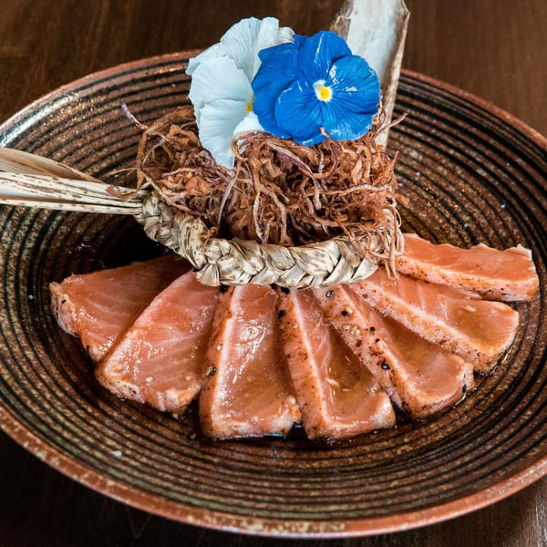 Seared salmon sashimi with crispy fried onions and edible blue and white flowers on a brown plate.