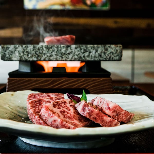 Marbled raw beef slices on a white plate with a hot stone grill and flames in the background.