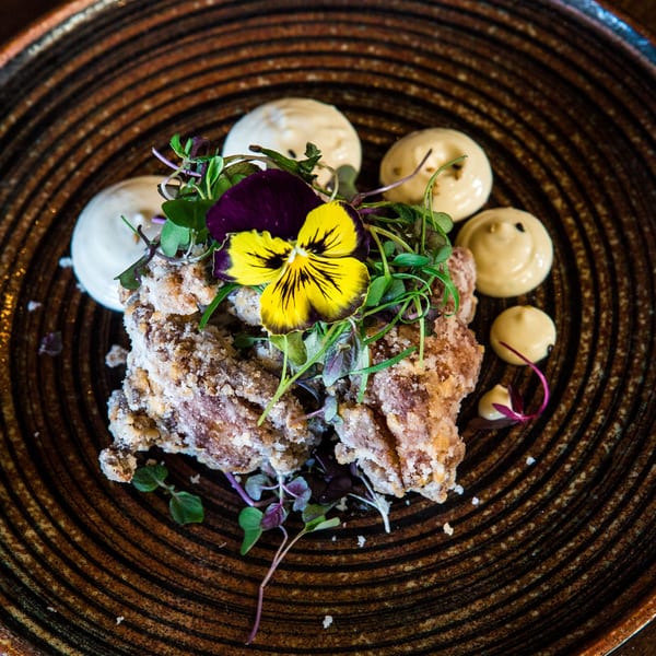 Crispy fried chicken karaage with microgreens, a yellow edible flower, and dollops of sauce on a dark plate.