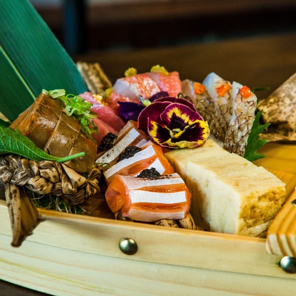 A wooden boat filled with various sushi, sashimi, and tamagoyaki, garnished with a purple flower.